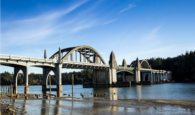The Siuslaw River Bridge in Florence, Oregon, on a bright day. There are wisps of clouds in the sky and the shore on the other side of the river is lined with trees.