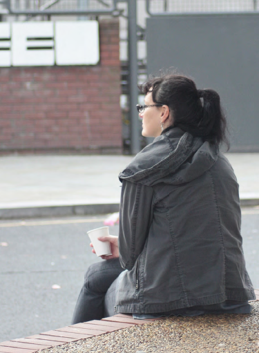 A young adult presenting as female, sitting on a cement step alone outside, with back and side facing the camera so we see a profile view