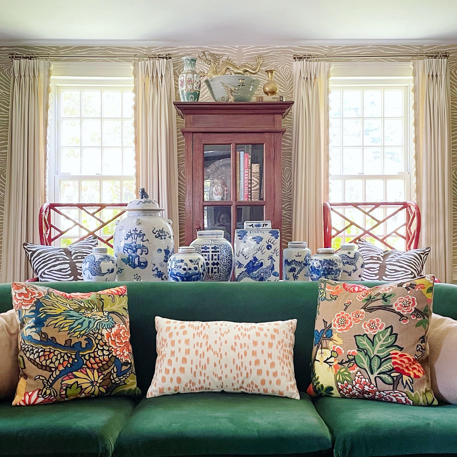 As you enter the living room, you are immediately drawn to the beautiful chinoiserie chairs placed by the windows. The chairs are exquisitely designed with red color, and unique pattern. A plush green sofa sits in the center of the room, creating a cozy seating area for you to sink into.The two large windows in the room allow plenty of natural light to filter in, creating a warm and inviting atmosphere. The windows are framed by flowing neutral color curtains that complement the blue and white vases displayed on the window sills. The vases are of various sizes and shapes, each unique and adding to the overall elegance of the room.The chinoiserie element is continued with the throw pillows on the sofa, each featuring a different design of flowers and dragon. A tall cabinet sits in the middle of the windows, its dark wood contrasting beautifully with the lightness of the room. The cabinet is adorned with more vases and other decorative items, adding to the room's charm.