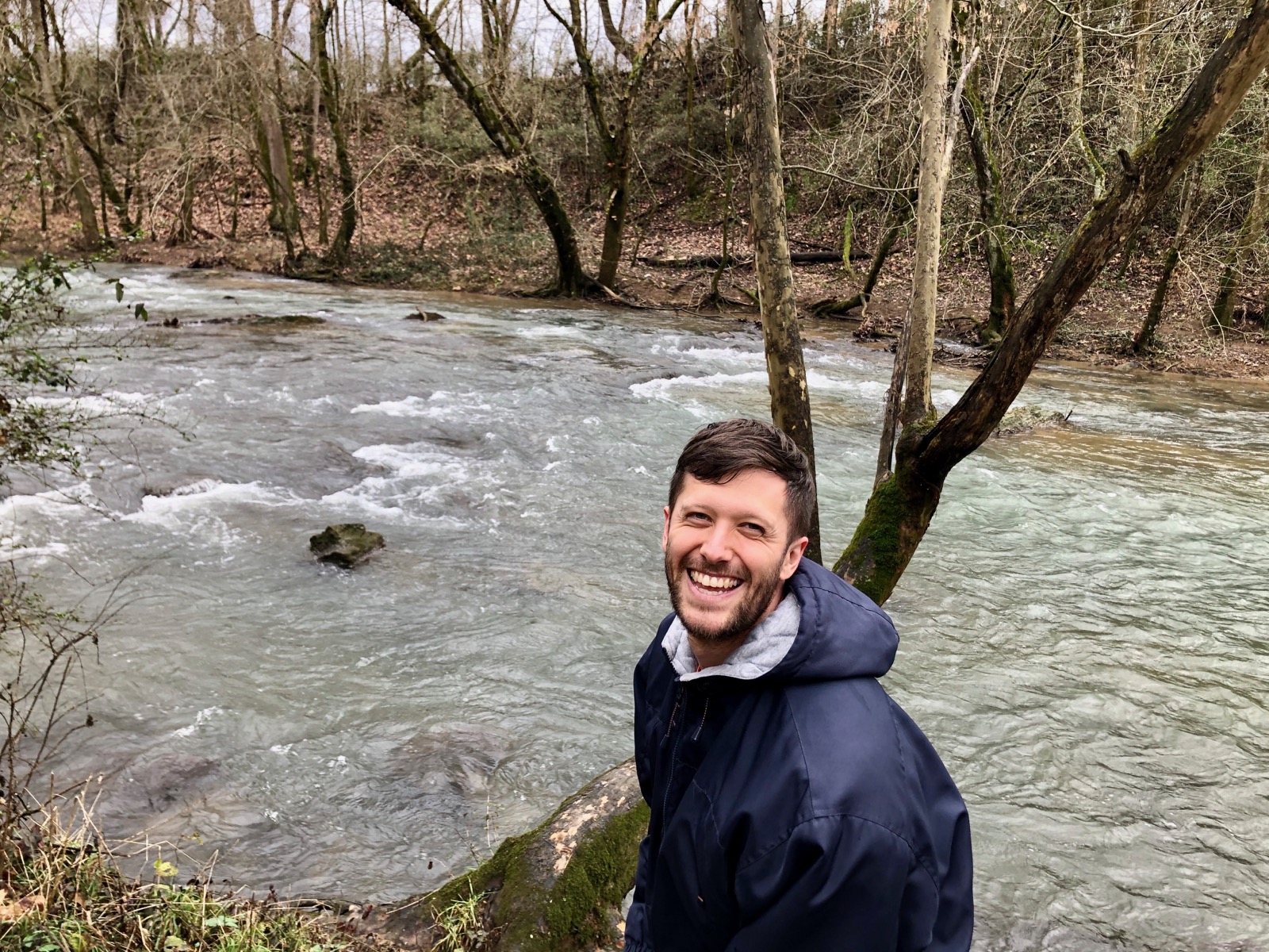 A man with brown hair standing in front of a flowing river laughs into the camera