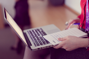 A MacBook laptop sitting on a person's lap. That person is also holding a notepad next to the laptop.  