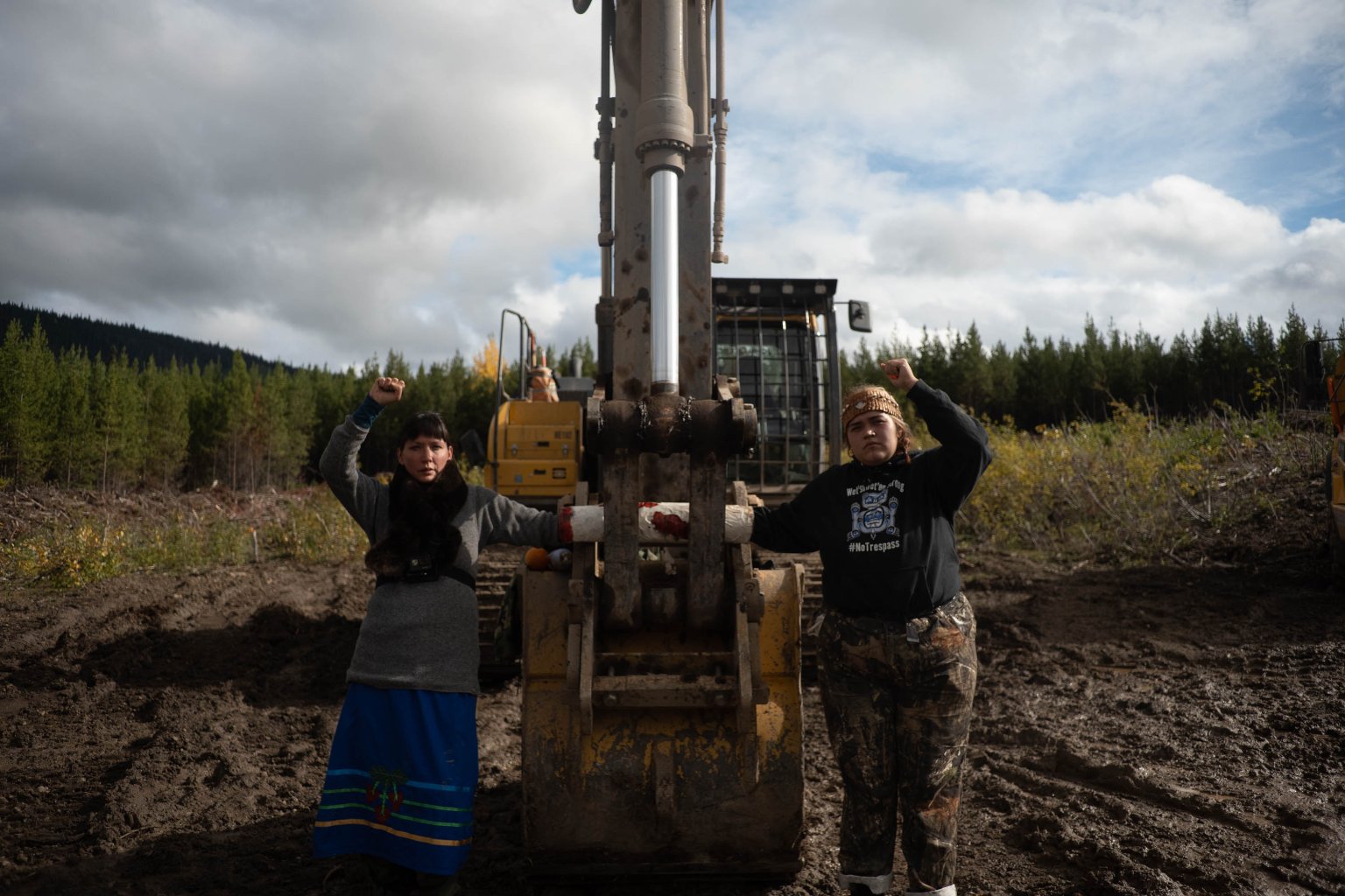 2 indigenous women stand and give a fist salute. They are in an arm lock attached to a mechanical digger in an area that has recently been cleared of vegetation.