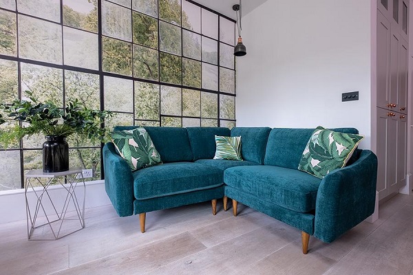 Để mang tới không khí thiên nhiên hòa vào phòng khách nhỏ, hãy chọn một chiếc ghế sofa góc V xanh cổ vịt có chân bằng gỗ kết hợp gối trang trí họa tiết lá cây