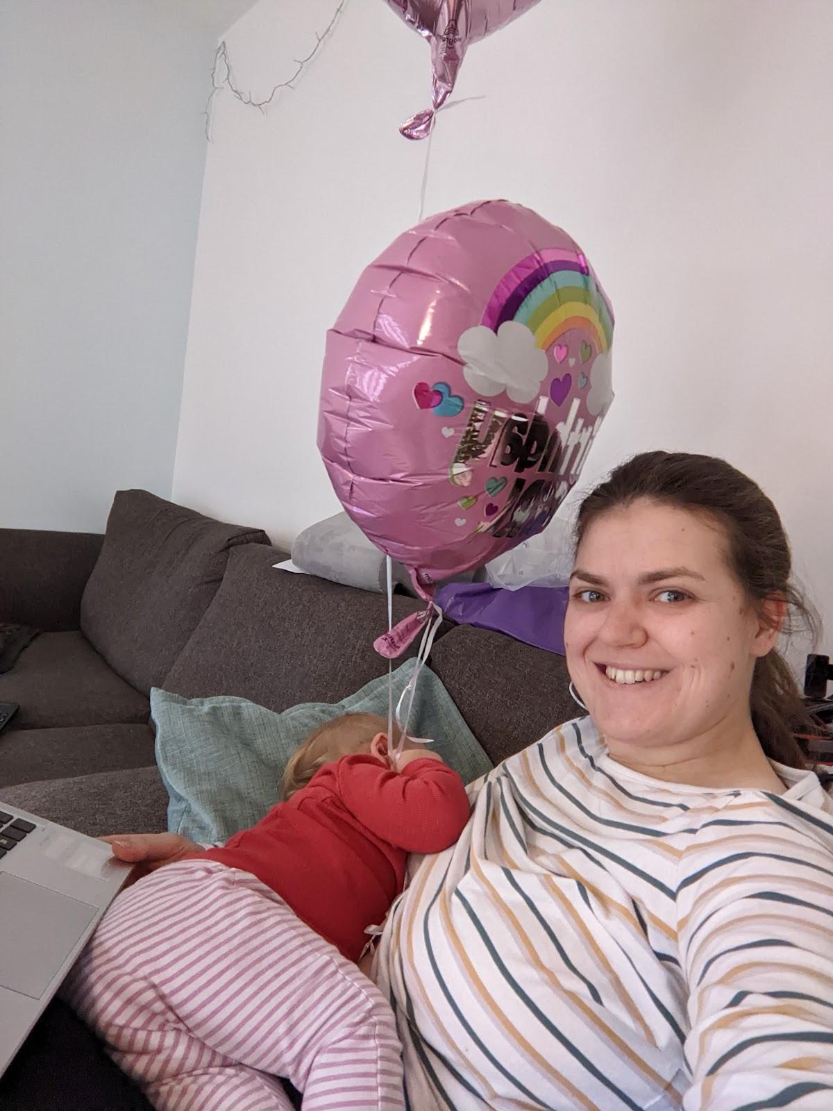 Sitting on my sofa, supporting my daughter with one arm, typing with the other and balancing both her and my laptop on my lap. My daughter is holding the strings of two helium birthday balloons.