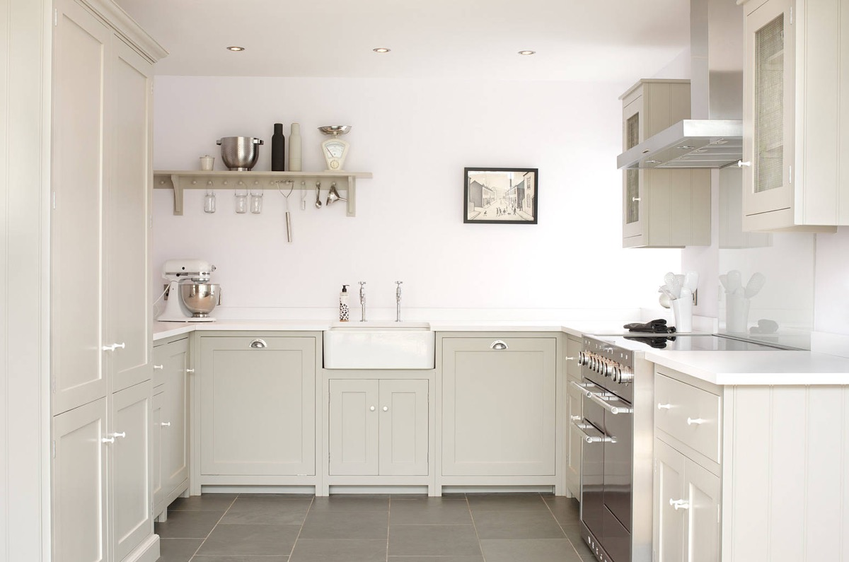 small u-shaped kitchen with light beige shaker cabinets. the far wall is left bare except for one open shelf, and white paint is substituted for kitchen backsplash