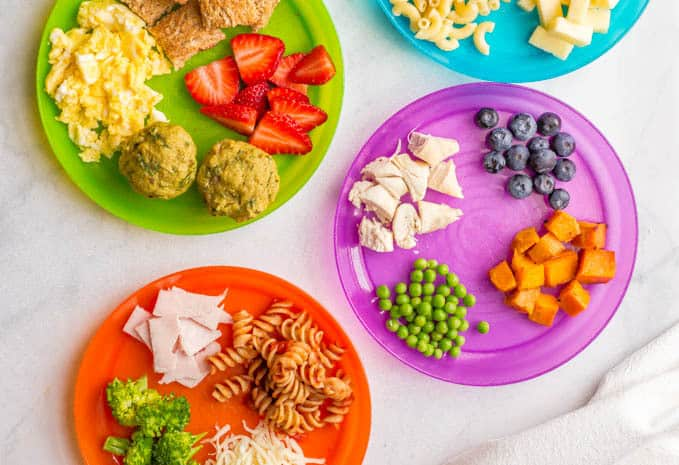 scrambled eggs, strawberries and toast served on a green plate; diced chicken, blueberries, sweet potatoes and peas served on a purple plate; pasta, slices of turkey and broccoli served on an orange plate