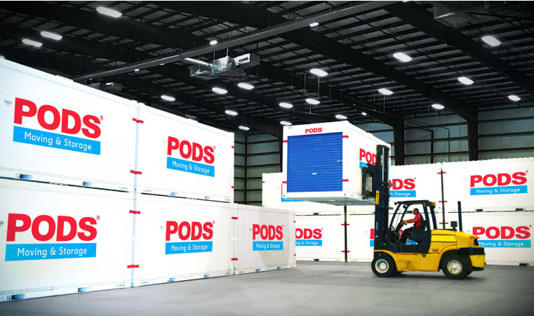 Eleven PODS portable storage containers are neatly stacked in a PODS Storage Center. A PODS employee is using a forklift to position another container in the group.