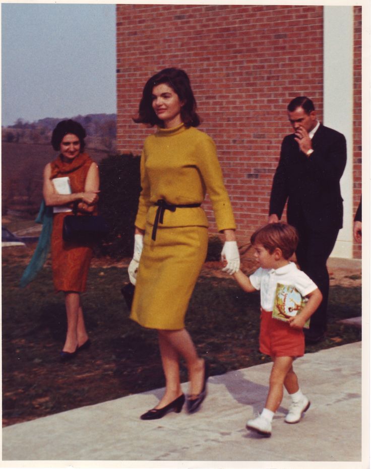 Jacqueline Kennedy, com um conjunto inteiro mostarda, acompanhada de seu filho pequeno em uma rua. Foto tirada em 1963. 
