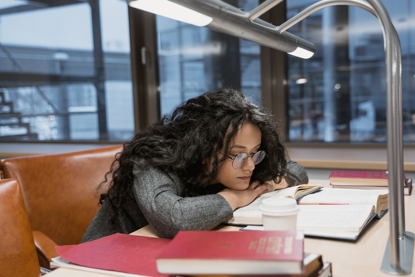 A teen girl wearing glasses is reading over multiple textbooks with a cup of coffee on the table. 