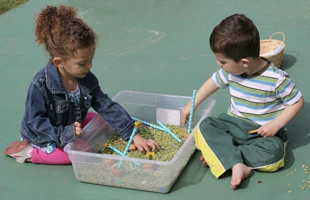 Two children, a boy and a girl playing with split beans poured in a plastic container with plastic straws and sequin flowers