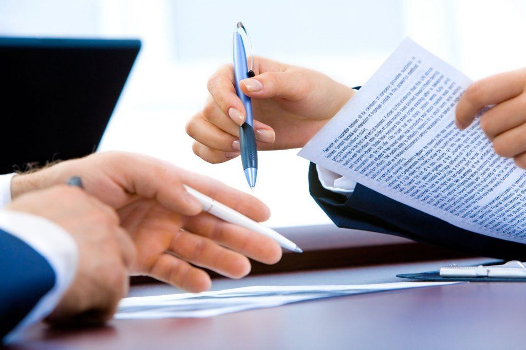 A slew of papers on a table with two people going over them while in an office setting.