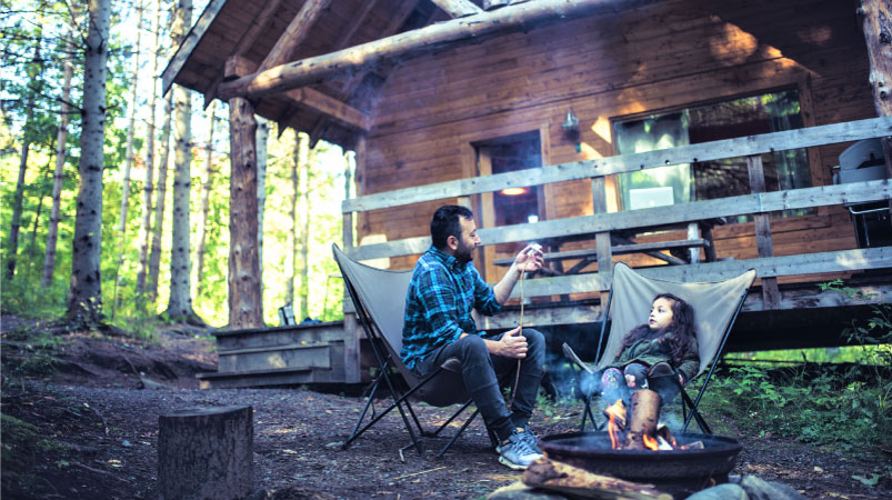 A father and daughter sit in front of a cabin in the woods sharing a bonding moment by roasting marshmallows over a campfire.
