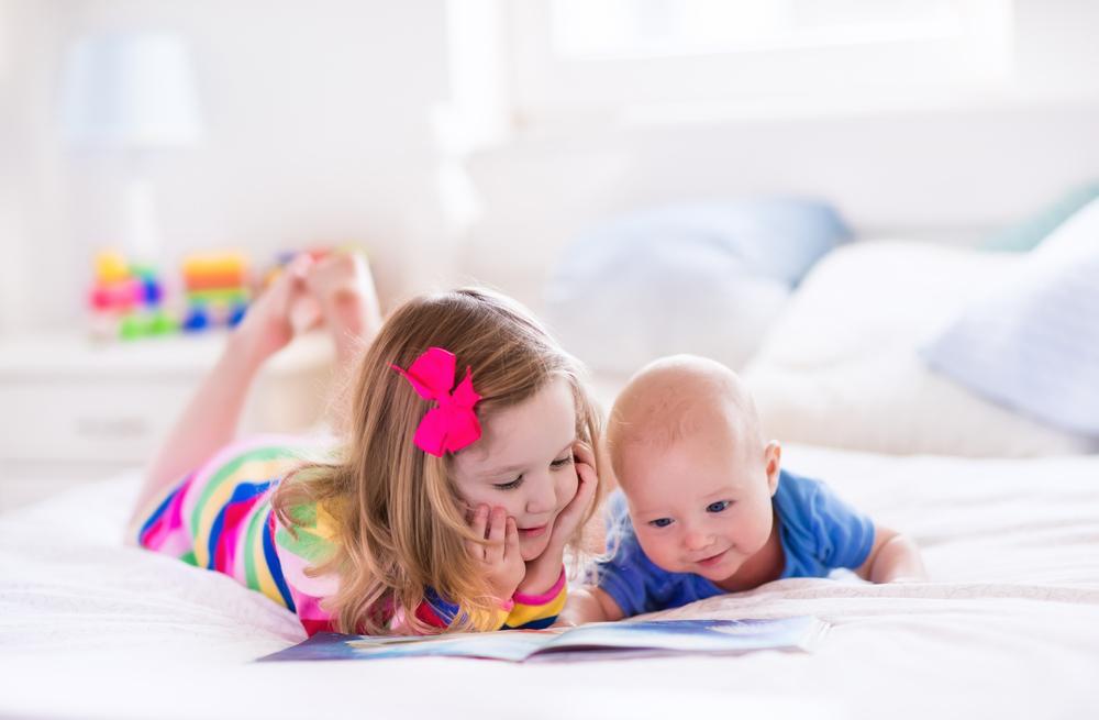 Funny happy toddler girl reading a book and playing with newborn baby boy in bed. Kids play at home. White nursery. Child in sunny bedroom. Children read and study. Interior for baby and young kid.