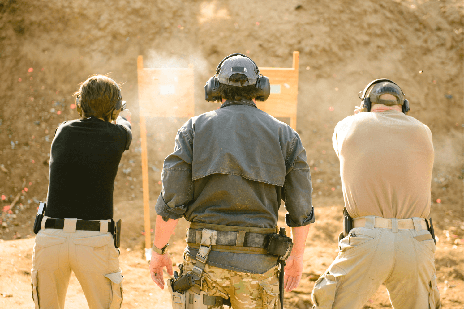 Two men and an instructor at an outdoor shooting range are firing rounds from their weapons.