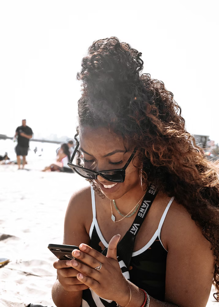 A woman with curly brown hair checking her phone on a sunny beach. 
