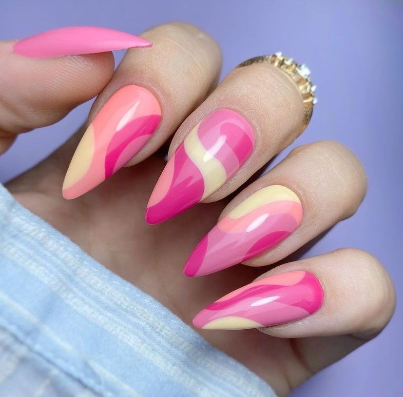 Mãos de uma pessoa com unhas no formato stiletto, que consiste em deixá-las alongadas e com as pontas bem marcadas. As unhas estão decoradas com esmalte rosa pink, rosa claro e amarelo. 