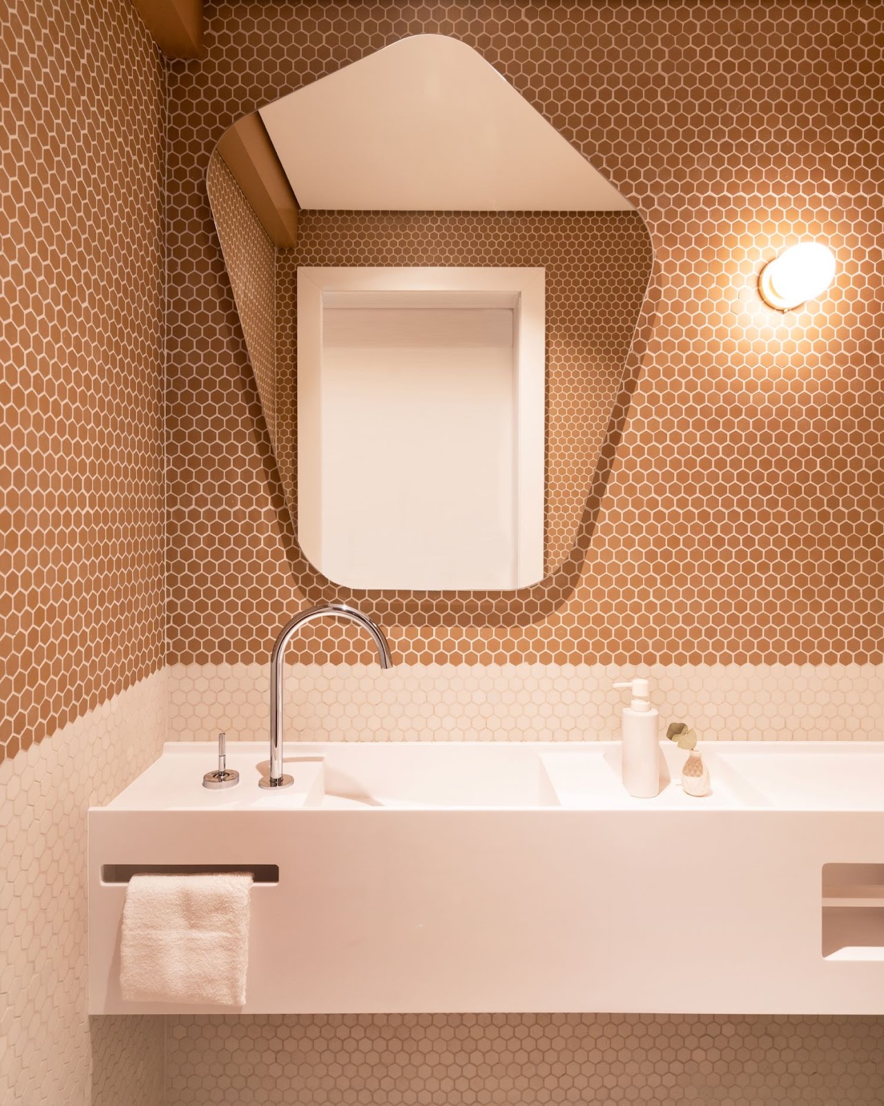 Banheiro com bancada e pia branca, parede revestida com pastilha em formato hexagonal rose e nude e espelho orgânico na parede.