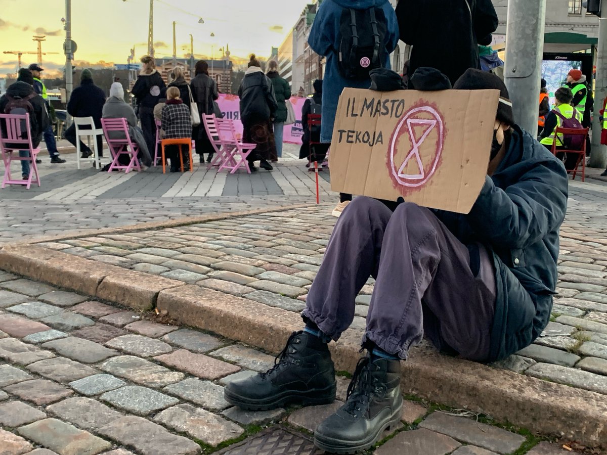 A rebel sits on a curb with an XR sign covering his face. Behind we see rebels sat in the road on pink chairs having a meeting