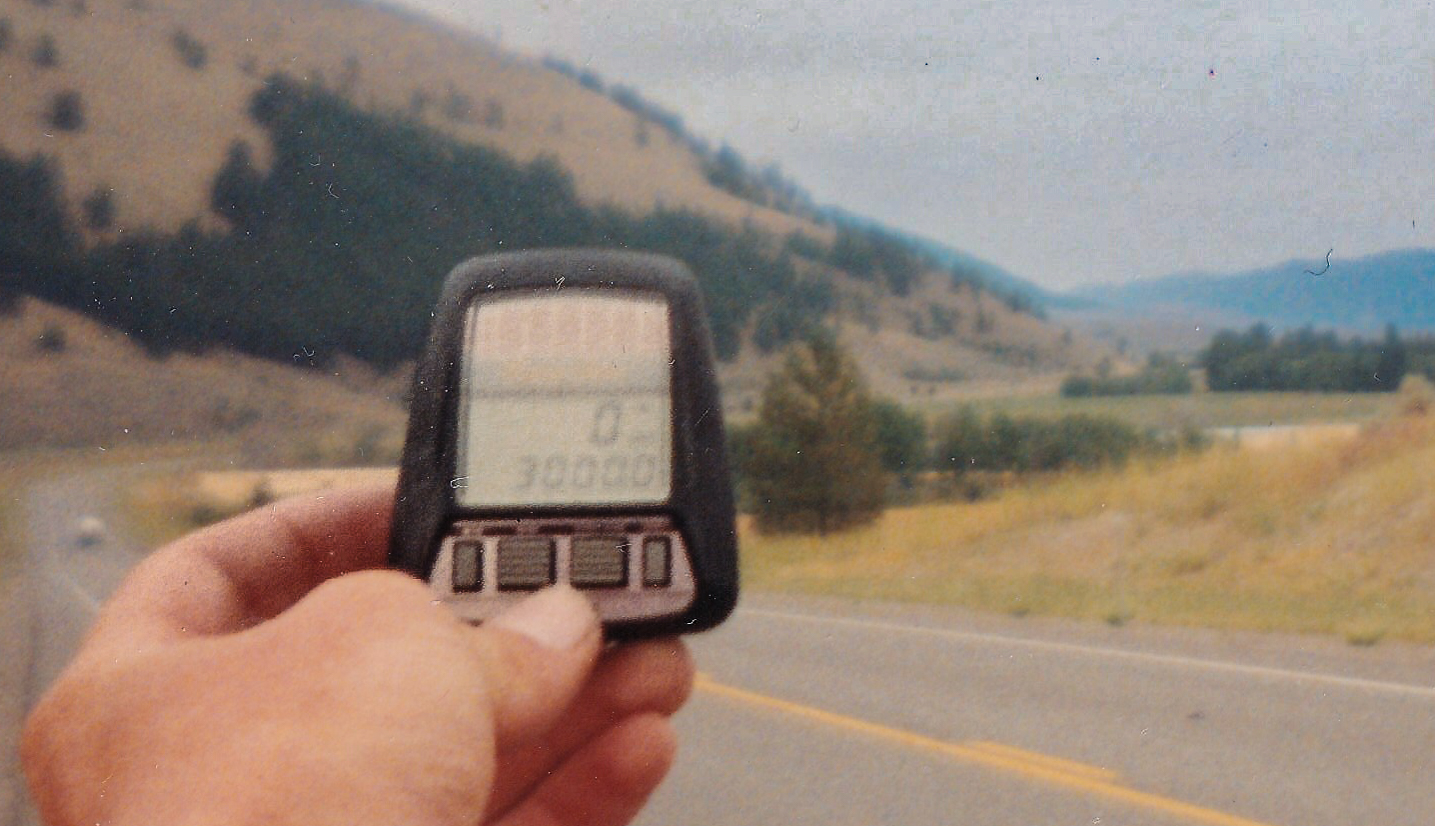 A hand holds a small bike odometer that reads 3,000 miles