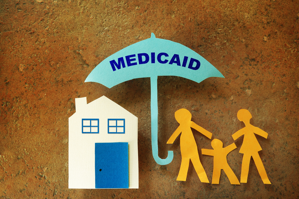 Paper cutouts of a family with a house, covered by an umbrella labeled 'Medicaid.'