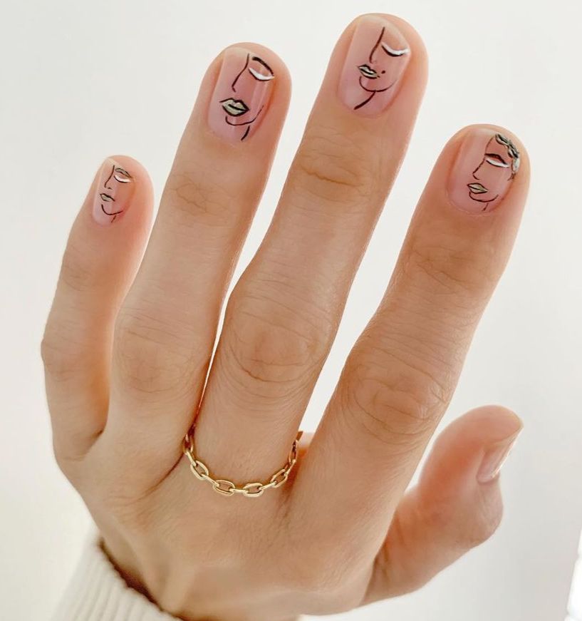 Mão de uma mulher com unhas decoradas de forma minimalista da artista Betina Goldstein, com traços de rostos em preto e detalhes prateados.