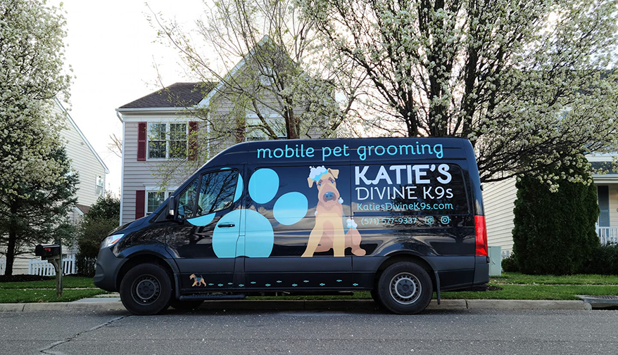 A vibrant mobile pet grooming van parked in front of a charming house. The van features a colorful ad poster on its side, showcasing a cheerful dog enjoying a refreshing bath. The ad prominently displays the contact details for Katie's Divine K9s, including a phone number and email address.