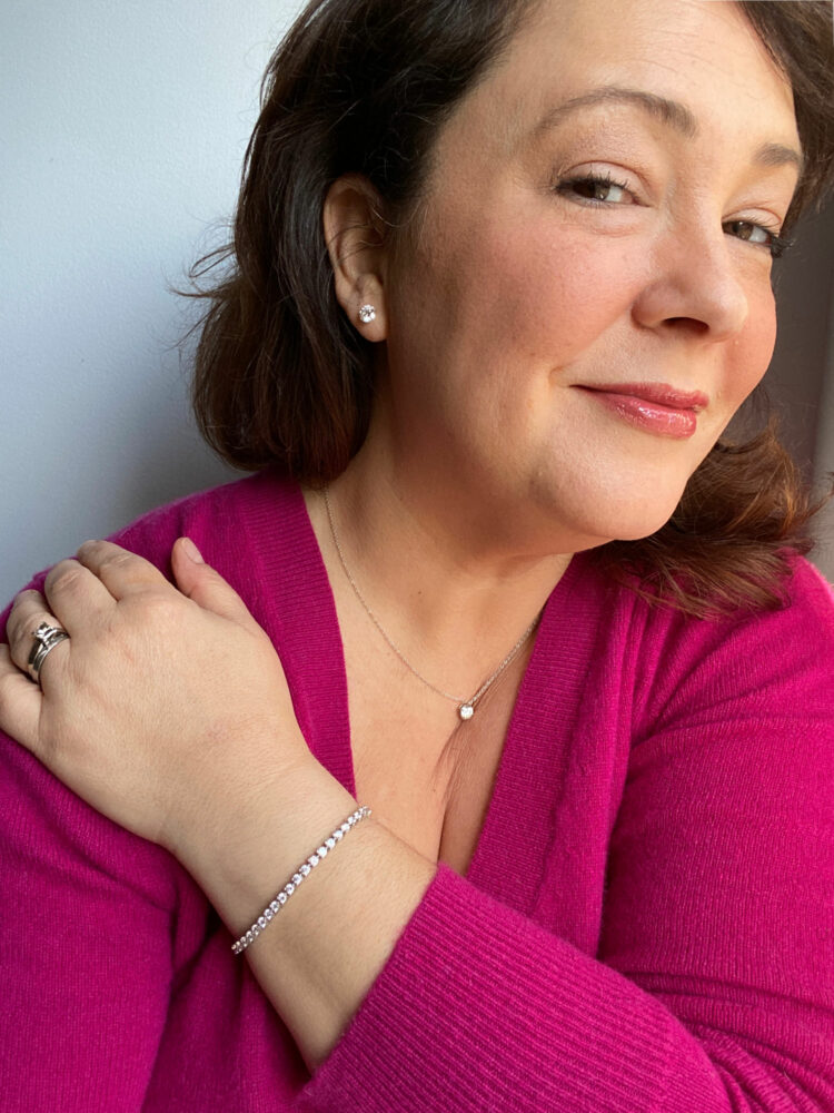 Alison Gary wearing a berry colored cashmere sweater and is looking at the camera with a slight smile. Her left arm is across her body with her hand on her right shoulder.