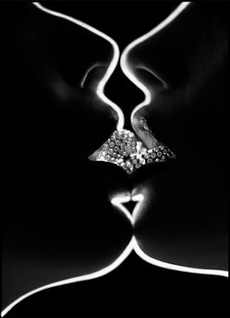 Silhueta do perfil dos rostos de duas modelos femininas que estão próximas inclinadas de frente para outra. As bocas ganham destaque e ambas carregam anéis de diamante que se encostam pelo beijo.