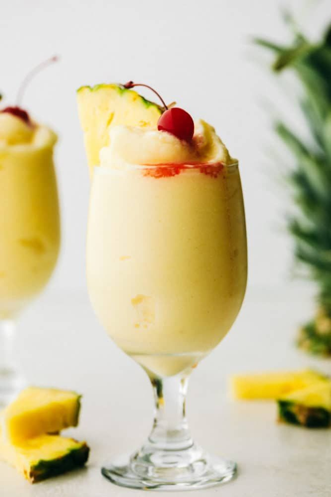 A imagem mostra um copo do drink pinã colada, decorado com folhas e um pedaço de abacaxi. Fora de foco, é possível perceber outros copos e rodelas de abacaxi. Também há uma cereja no topo do drink.