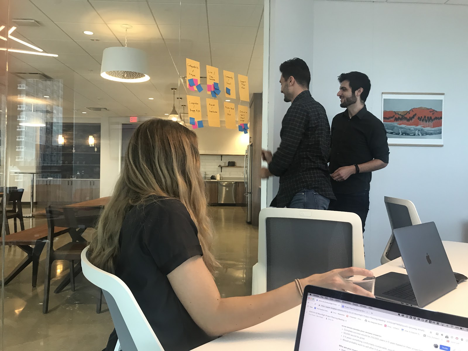 Three people Dot Voting by placing colored sticky notes on goals of their choice, posted on a glass wall