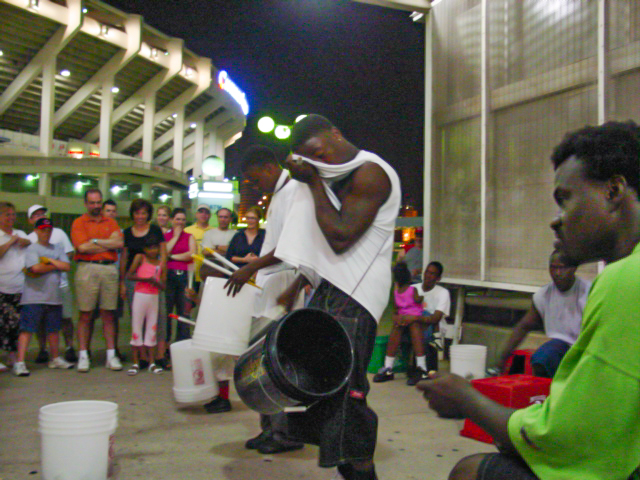 Man sits in the foreground wile four companions drum for a crowd at night outside a stadium.  
