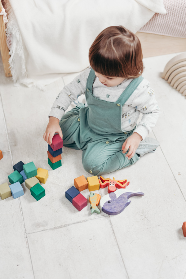 A child in a long-sleeved shirt and light green overalls sitting on the floor with toys and stacking blocks.