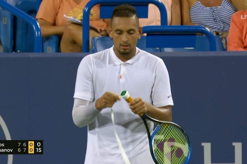 While standing on court, Kyrgios wraps a white grip around the handle of his racquet.