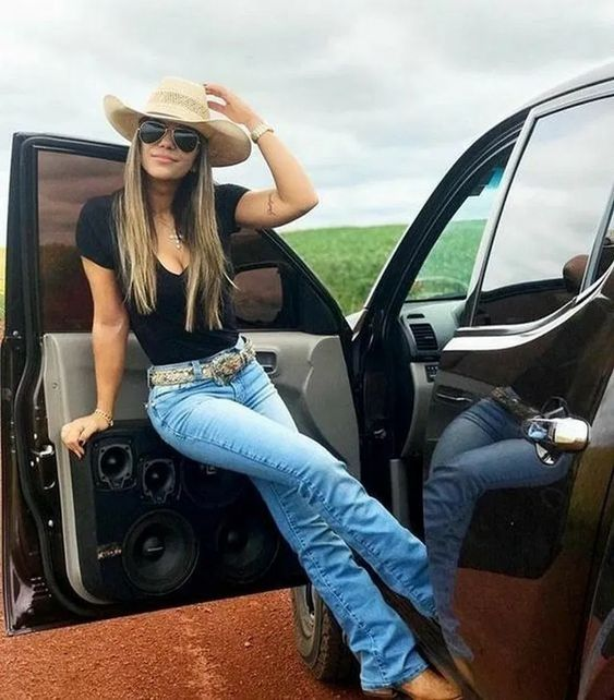 a lady in a pair of jeans, a black top, and a cowgirl hat by her car