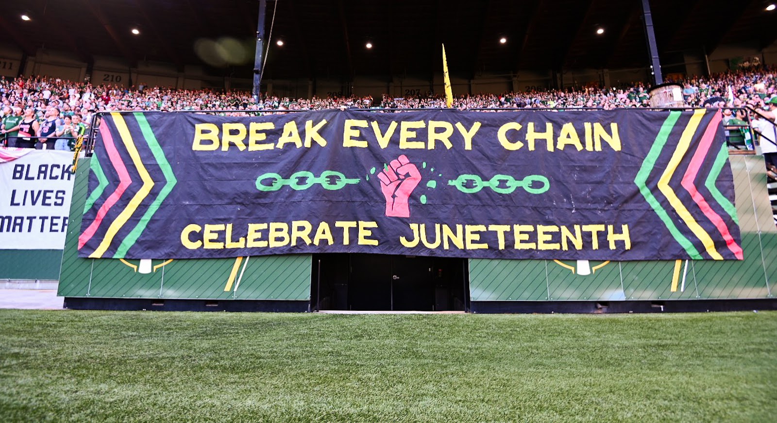 maintstage banner in Providence Park, black background with the words "BREAK EVERY CHAIN CELEBRATE JUNETEENTH" in yellow letters. There is a red fist going upwards through a green chain. Yellow, green, and red arrows on both sides of the banner pointing towards the message. 