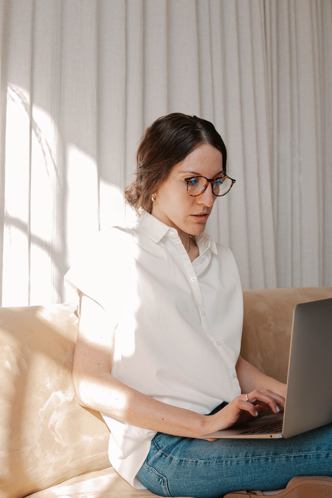 An image of a woman wearing glasses, sitting on a sofa, typing on a laptop computer.