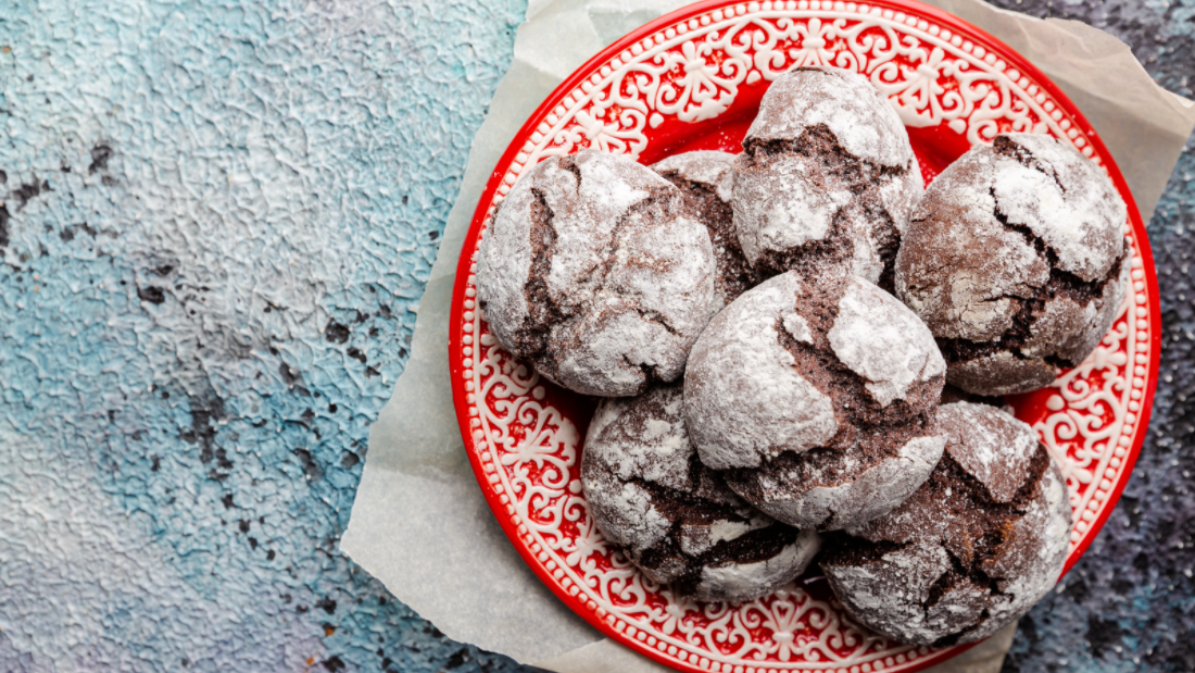 A tray of chocolate crinkle cookies arranged on a red holiday plate with a napkin under the plate on a marble countertap.