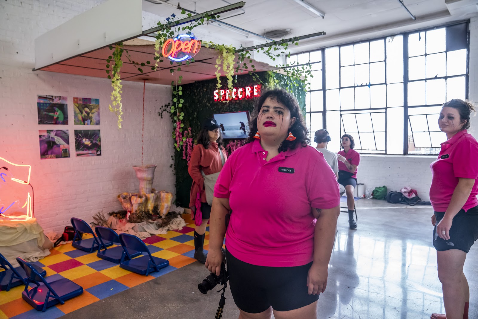 Image: A person wearing overdrawn lipstick and running mascara aloofly stands in front of the Spacore exhibition. They dangle a camera in their right hand. Photo courtesy of Colectivo Multipolar.