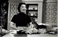 Woman with dark hair in a short-sleeved dark turtleneck shirt in front of a table with food spread out. A cupboard is in the background.