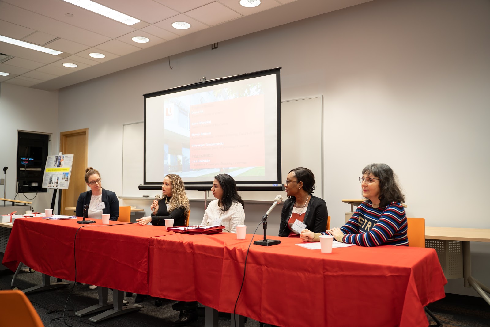 5 people are sitting at a table, 4 of which are panelists. The second person from the left is holding a microphone. 