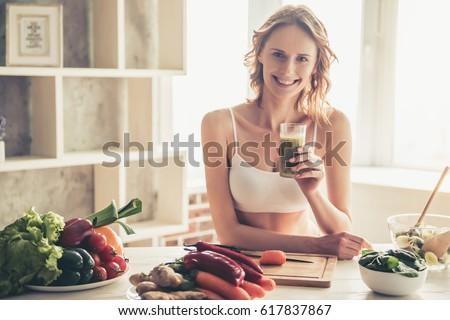 a smiling woman holding a glass of green juice and leaning on the counter full of vegetables
