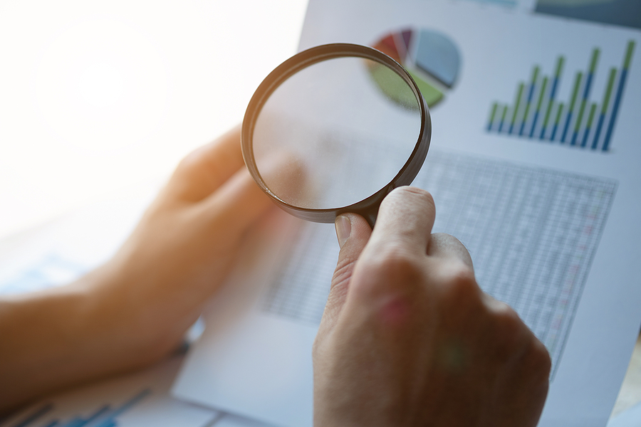 Close up of a business person examining budgets with financial documents in their left hand and a magnifying glass in their right. 