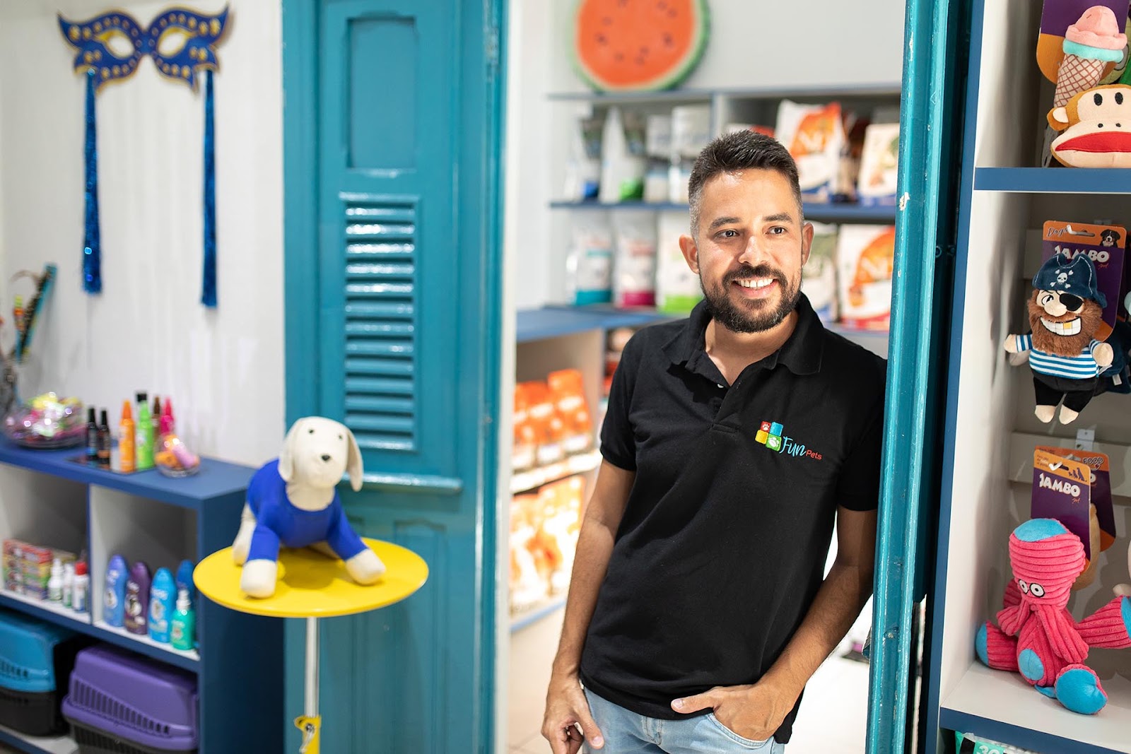 Homem moreno, de cabelo e barba grisalhos, veste camisa preta do pet shop Fun Pets. Ele está apoiado na porta e sorri olhando franquia de pet shop 
