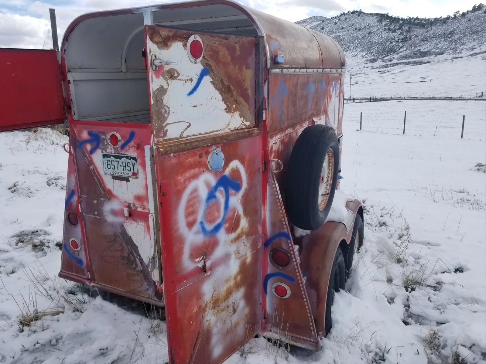 A rusted horse trailer spray-painted with graffiti rests in a snowy field at the base of the Colorado foothills.