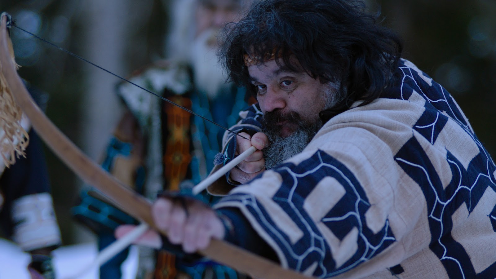 A screen still from Ainu Mosir, featuring a man holding a bow and arrow. He is notching the arrow on the bow, getting ready to let go.
