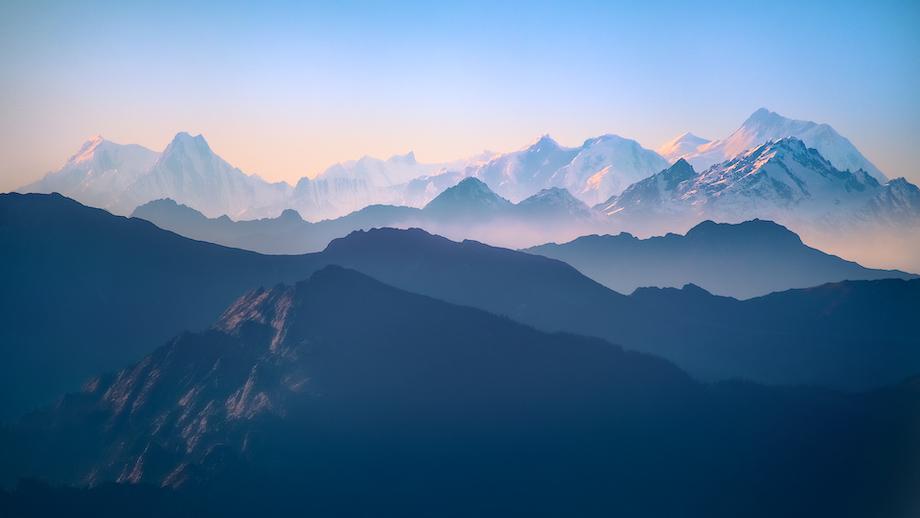 An image of a mountainous landscape, the low sun highlighting certain faces and peaks in pinks and oranges contrast with the dark blue hues of the rest of the range.