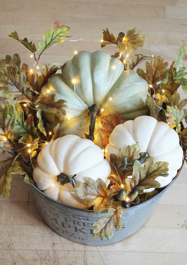 DIY pumpkin craft with three neutral colored pumpkins in a metal bucket. pumpkins are wrapped in fall foliage and christmas lights