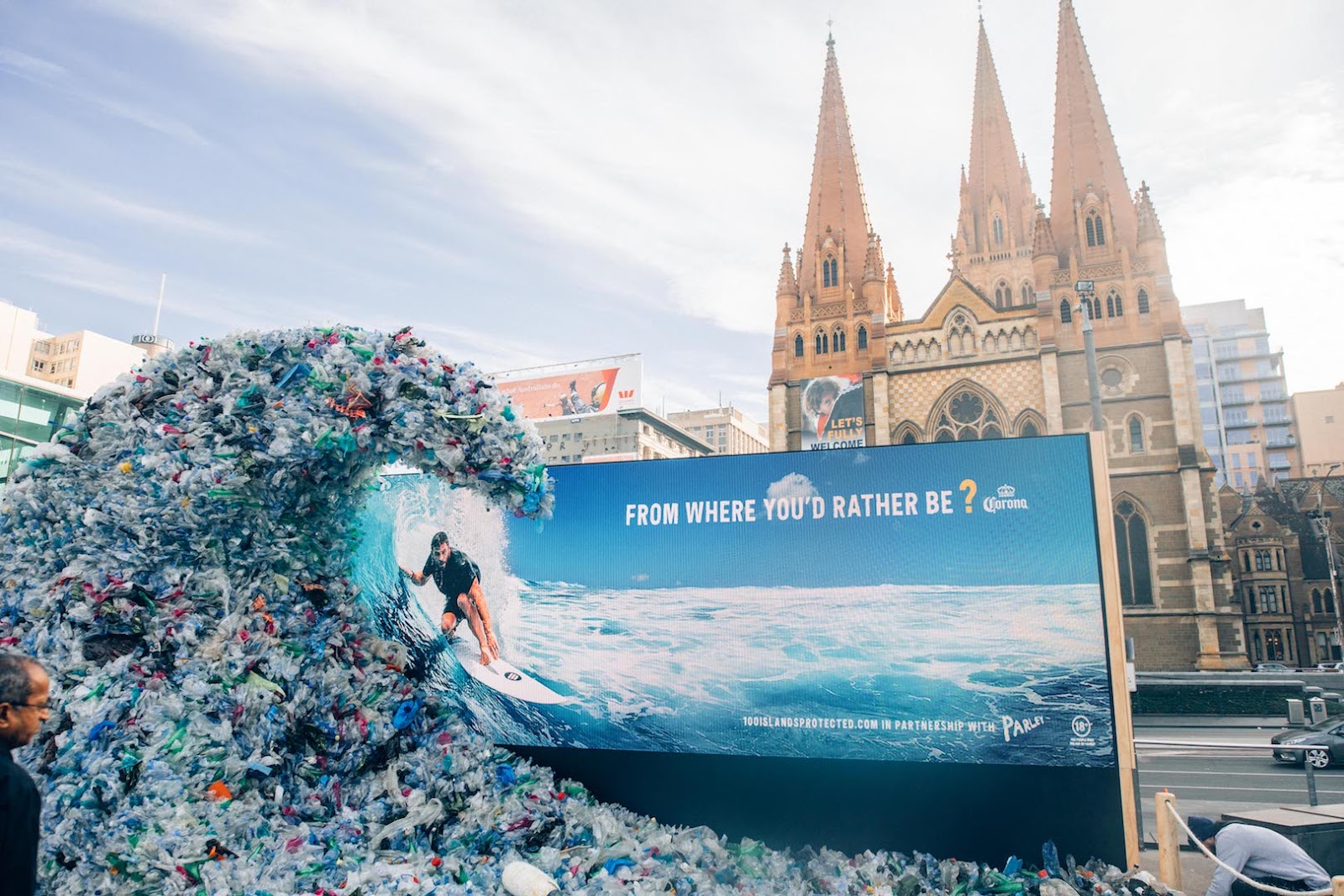 Giant plastic wave with a billboard behind, showing a man surfing in the real ocean and the text "From where you'd rather be?"