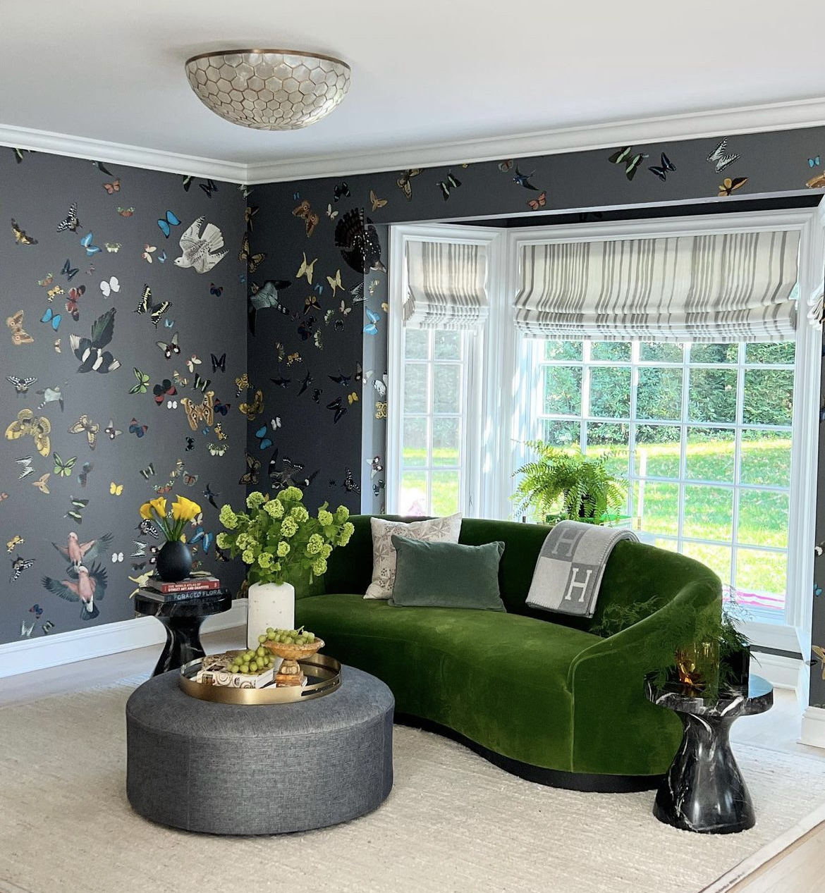 This stunning living room features a tufted curved sofa upholstered in a luxurious green velvet, a wall adorned with wallpaper featuring butterflies and birds, and a pair of modern side tables.