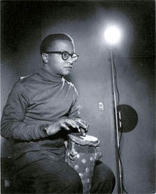 Billy Strayhorn, dressed in a dark turtleneck shirt, sits and plays a drum that he holds between his knees